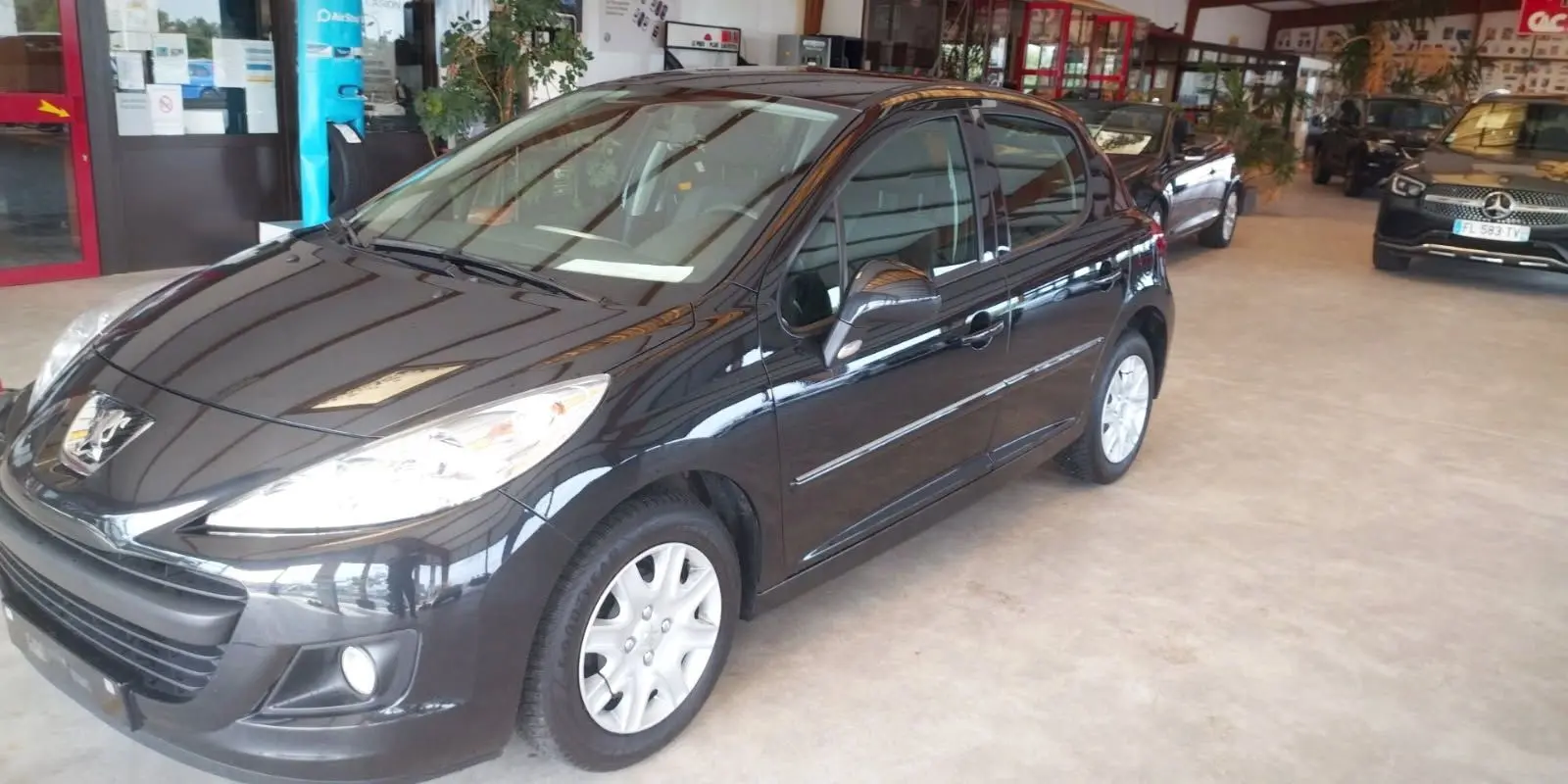 Peugeot 207+ noire vue en 3/4 avant droit dans un showroom, avec jantes argentées et phares allumés.