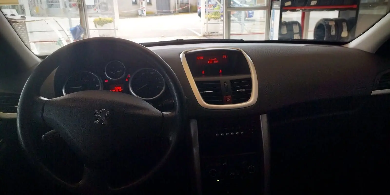 Intérieur de la Peugeot 207+ 2013 montrant le tableau de bord et le volant noir avec affichage numérique rouge.