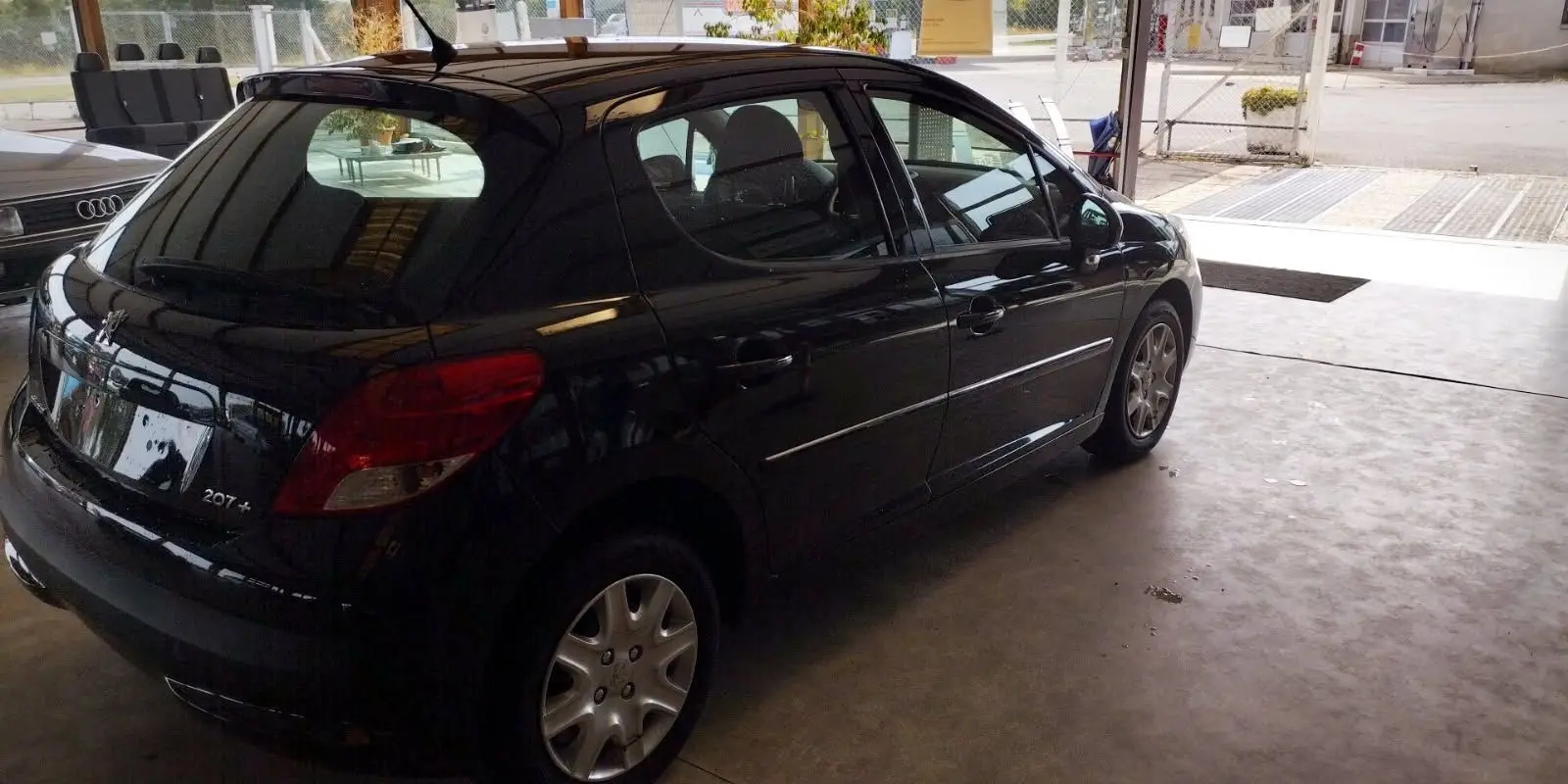 Peugeot 207+ noire vue de trois quarts arrière côté droit, stationnée dans un garage lumineux.