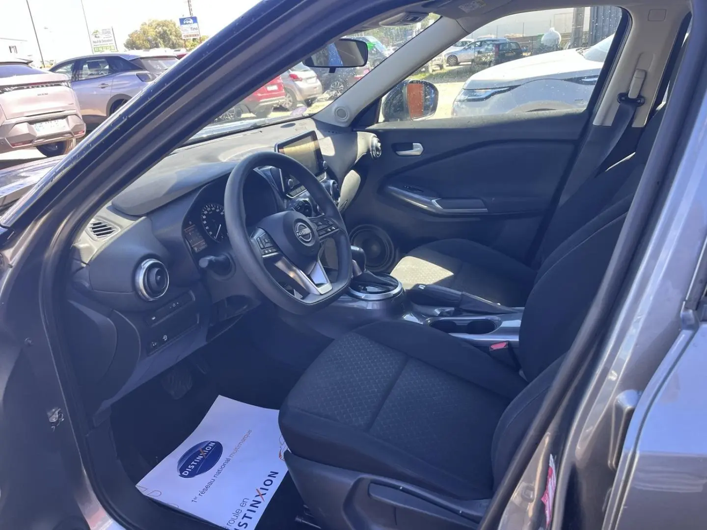 Vue intérieure côté conducteur du Nissan Juke 2023 gris argile, montrant le volant multifonction et les sièges en tissu noir.