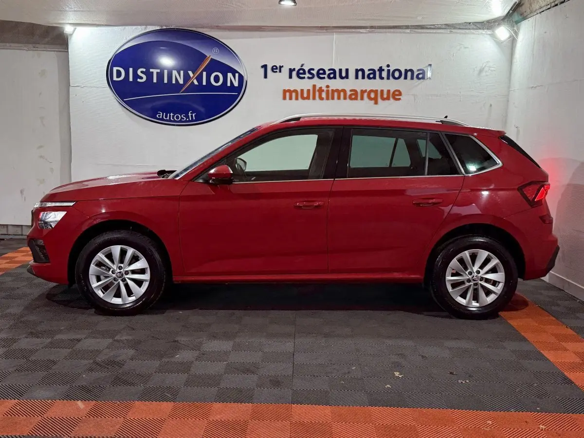 Vue latérale gauche d'un Skoda Kamiq rouge 2024 garé en intérieur avec jantes alliage et rails de toit noirs.