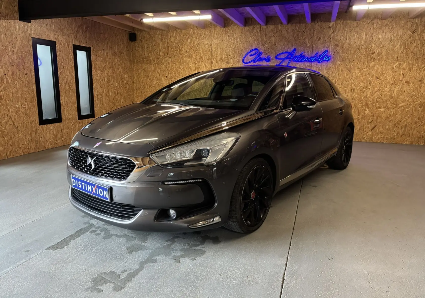 DS5 gris 3/4 avant droit en intérieur, avec jantes noires et calandre distinctive DS sous néon bleu.