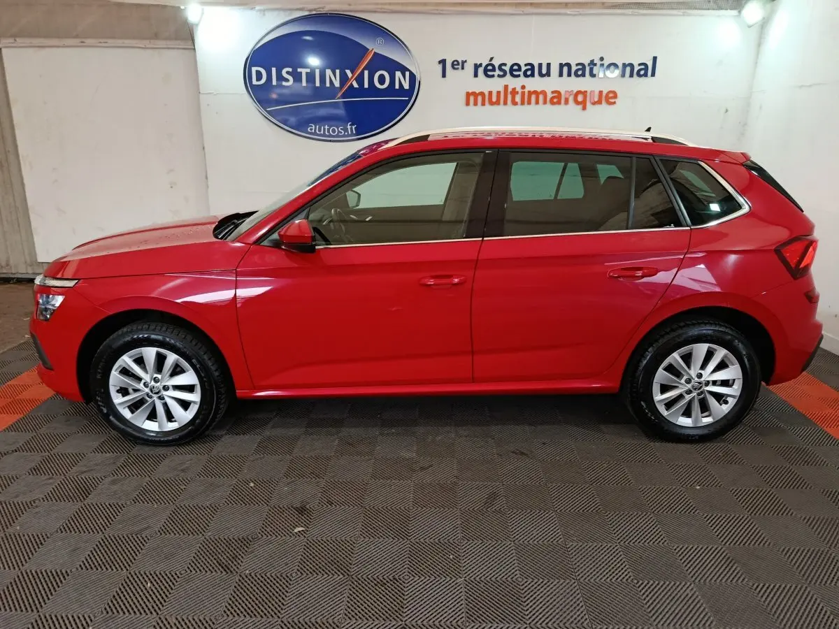 Vue latérale droite d'un Skoda Kamiq rouge 2024 avec jantes alliage et rails de toit noirs en intérieur showroom.