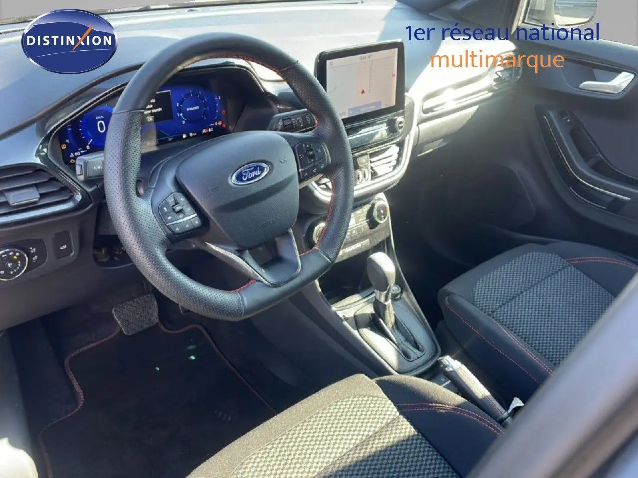 Vue intérieure côté conducteur du Ford Puma 2023, volant cuir perforé, tableau de bord numérique et sellerie tissu noir avec surpiqûres rouges.