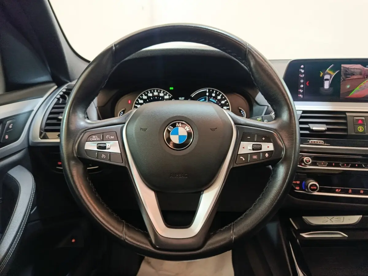 Vue intérieure centrée sur le volant cuir noir BMW X3 avec tableau de bord et écran multimédia en arrière-plan.