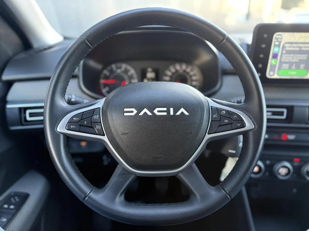 Vue rapprochée du volant noir du Dacia Jogger 2025 avec commandes intégrées et tableau de bord flou en arrière-plan.
