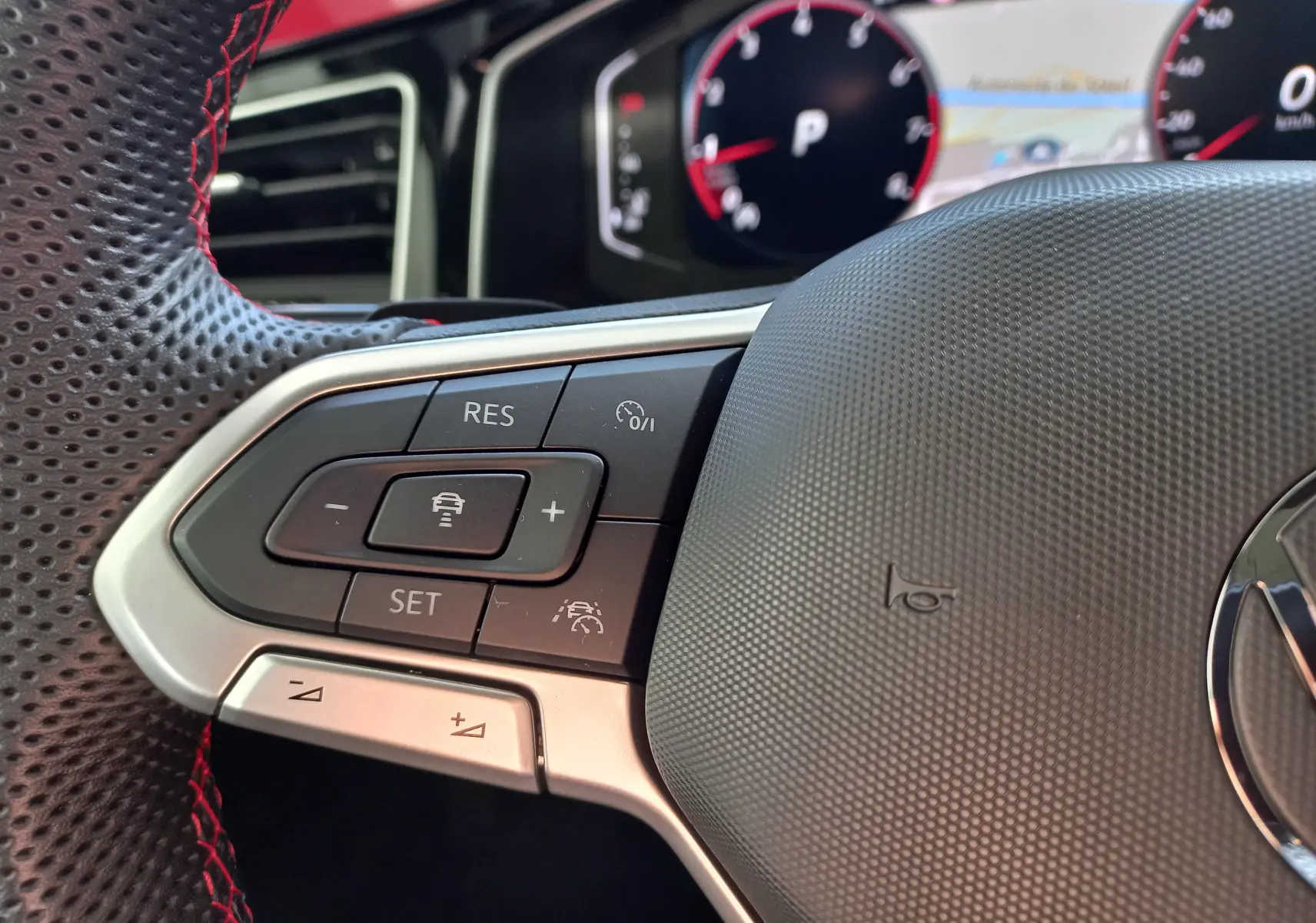 Gros plan sur les commandes à gauche du volant perforé avec surpiqûres rouges du Volkswagen Polo GTI blanc, tableau de bord numérique en arrière-plan.