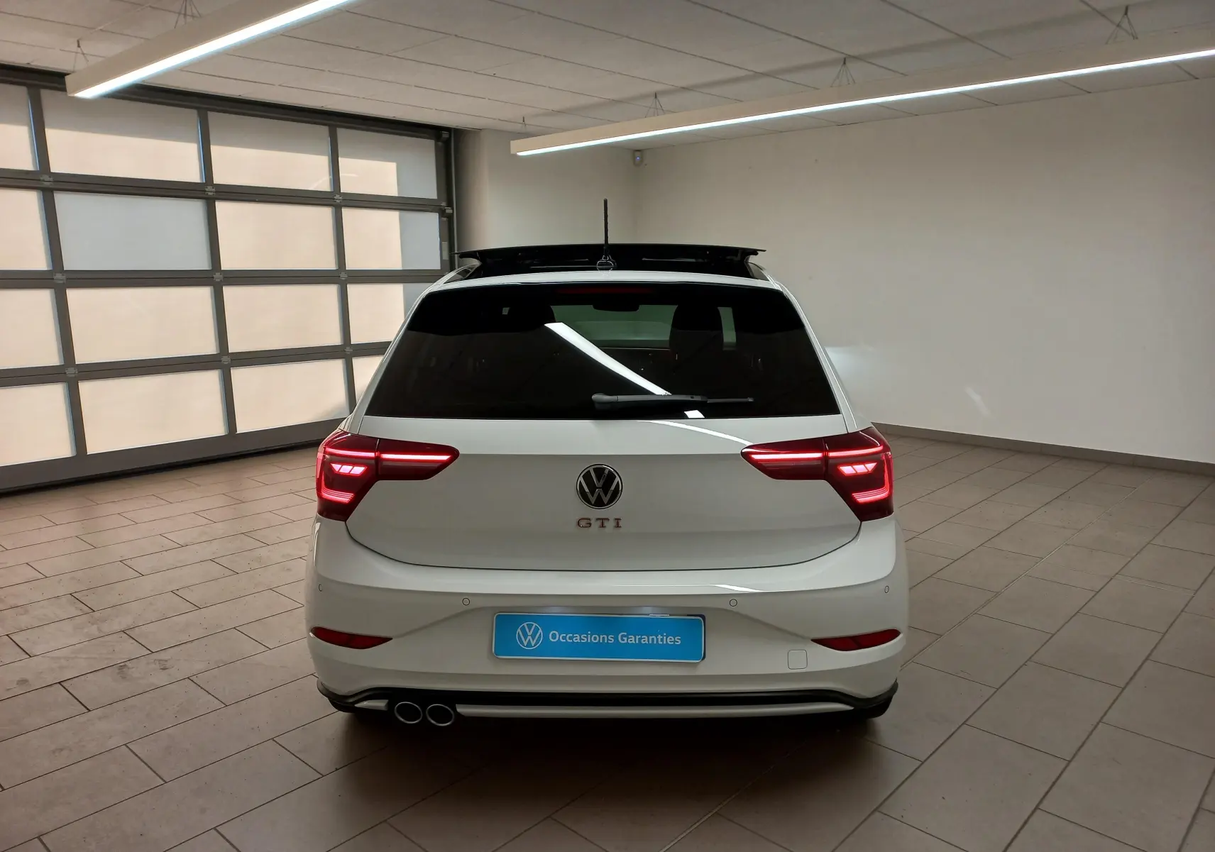 Vue arrière d'une Volkswagen Polo GTI blanche avec toit ouvrant noir et feux arrière allumés en intérieur showroom.