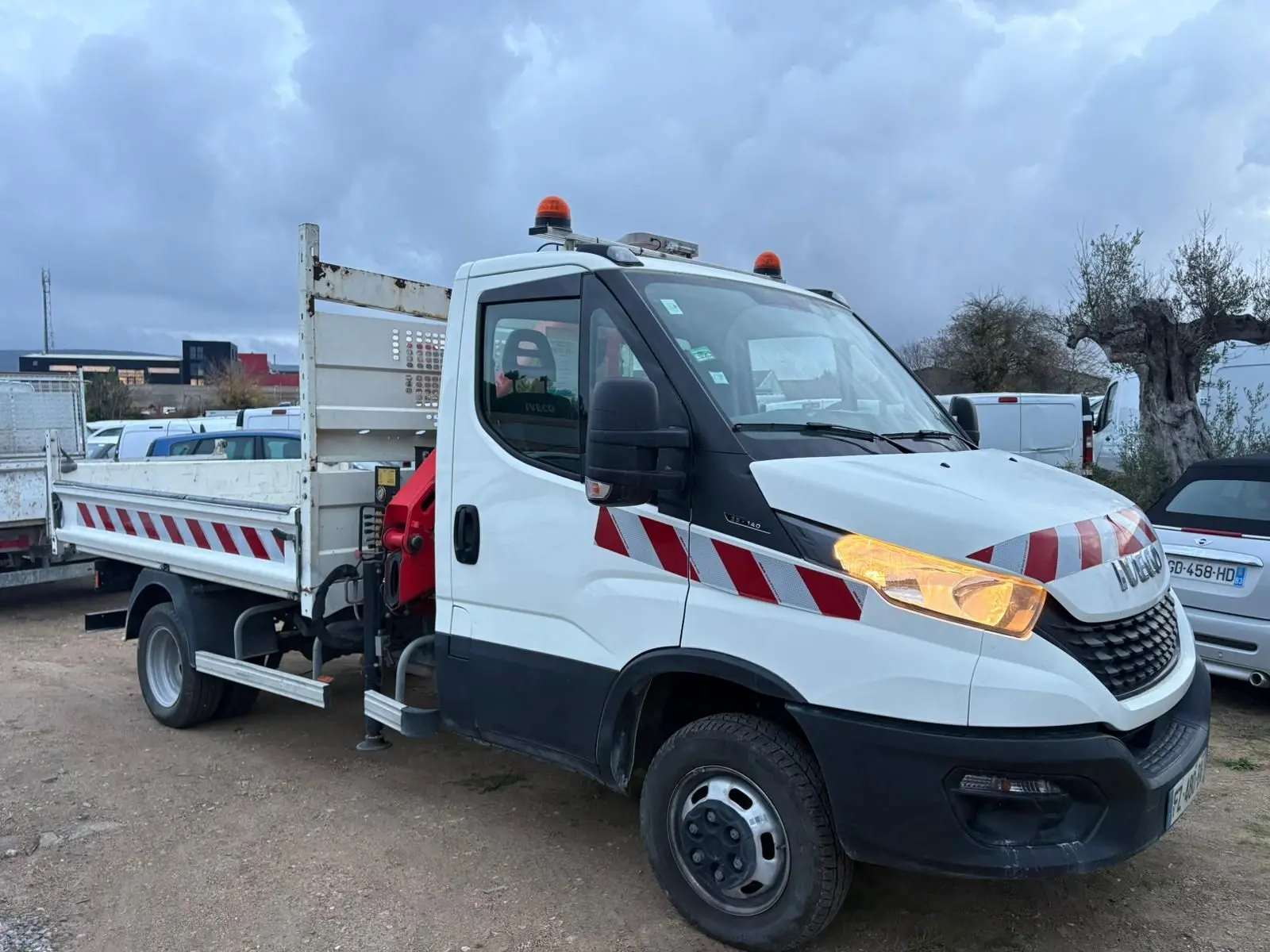 Vue 3/4 avant droite d’un Iveco Daily blanc avec benne, grue rouge Palfinger et bandes rouges réfléchissantes.