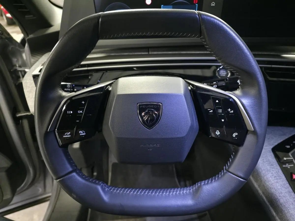 Volant compact en cuir noir avec logo Peugeot au centre, vue frontale rapprochée de l'habitacle gris chiné.