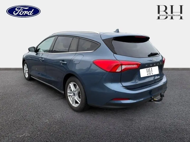 Ford Focus SW 1.5 EcoBlue bleu azur métallisé vue 3/4 arrière droit avec barres de toit et attelage.