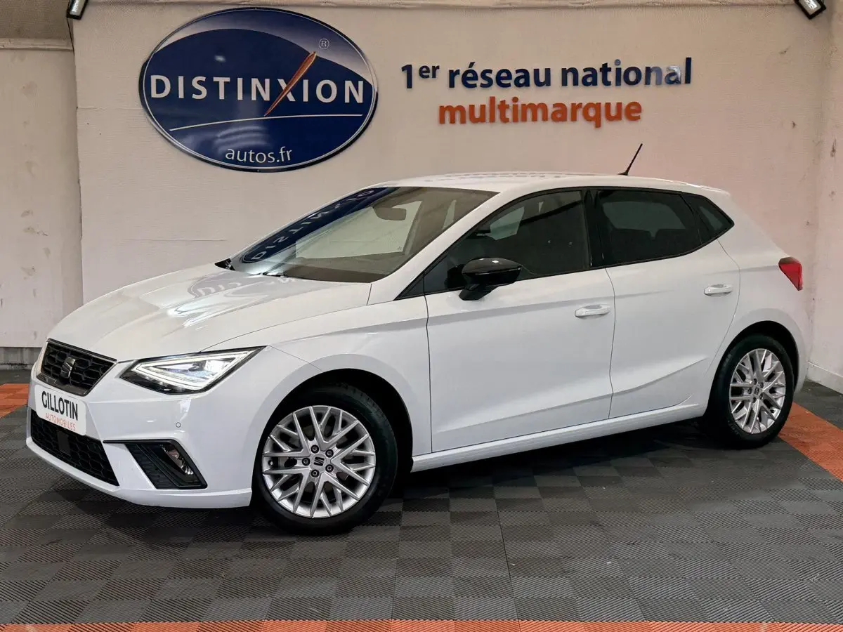 Vue 3/4 avant gauche d'une SEAT Ibiza 1.0 TSI 110 FR blanche avec jantes alliage et phares LED allumés.