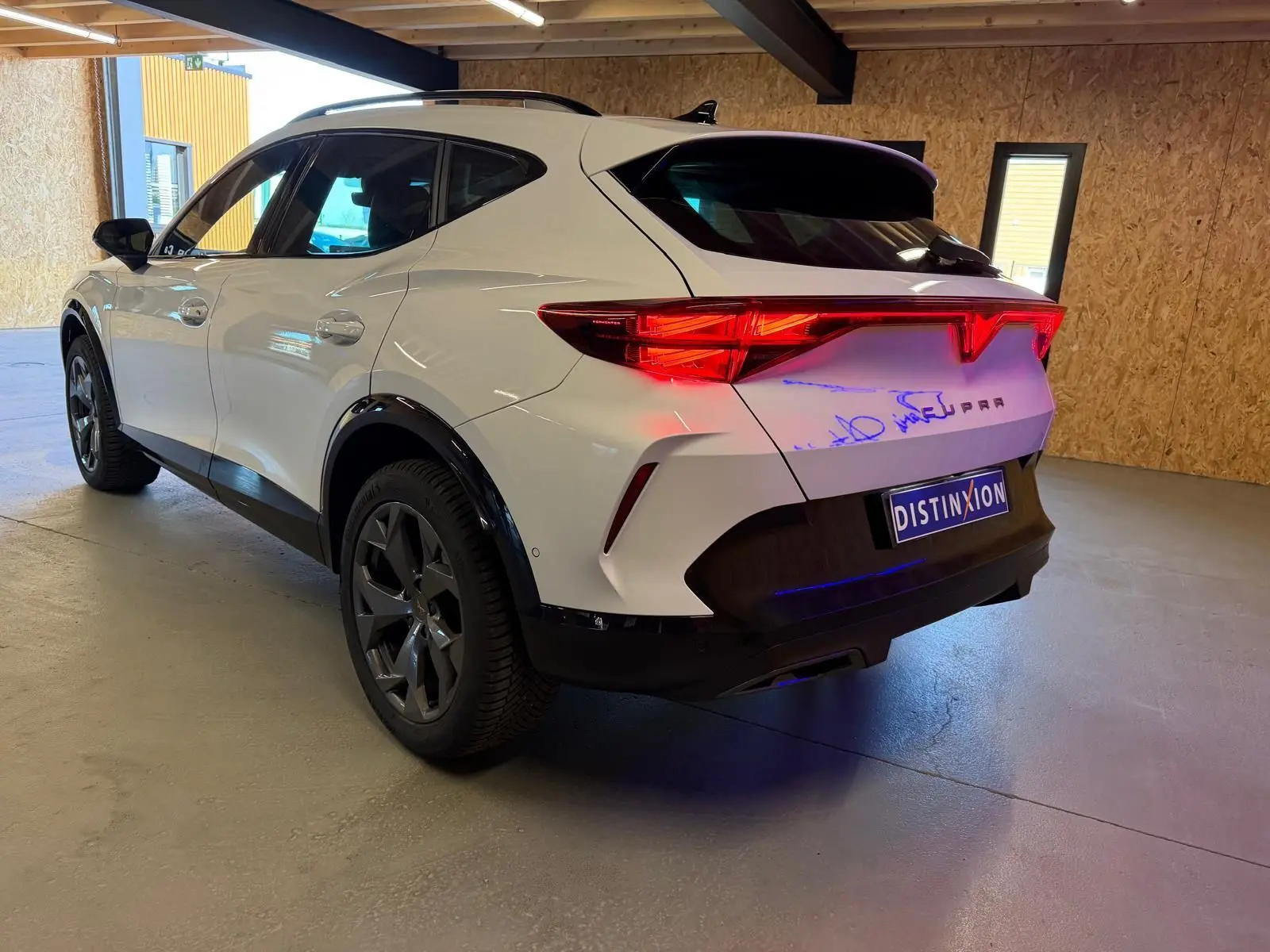 Vue 3/4 arrière droite d'une CUPRA Formentor blanche avec feux arrière allumés dans un garage.