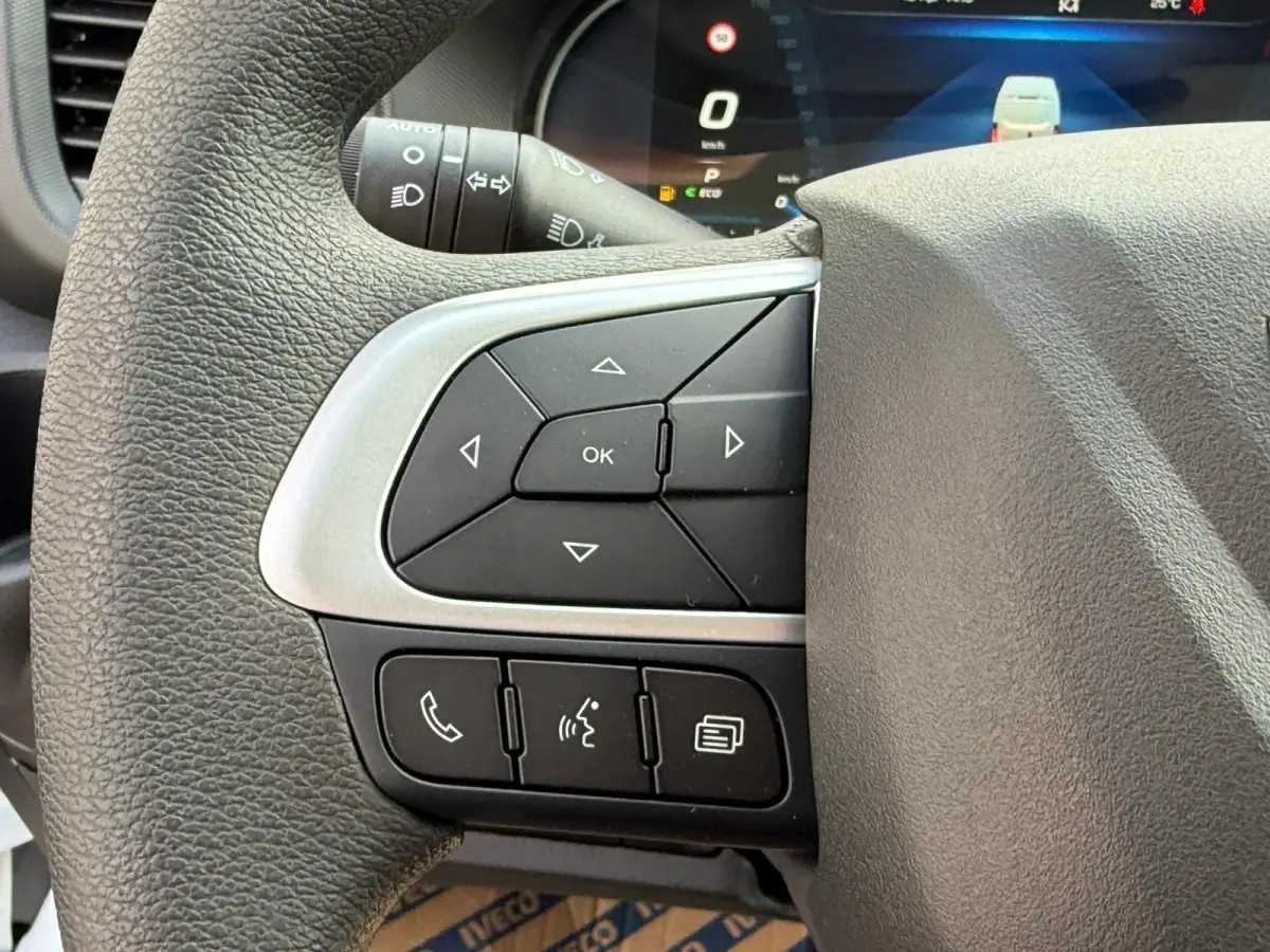 Gros plan sur les commandes gauche du volant noir du Iveco Daily blanc, avec affichage digital en arrière-plan.