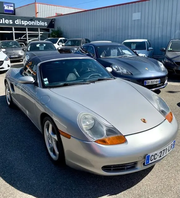 Porsche Boxster 986 gris vue 3/4 avant droit, capote noire et feux avant caractéristiques orange, stationnée en extérieur.