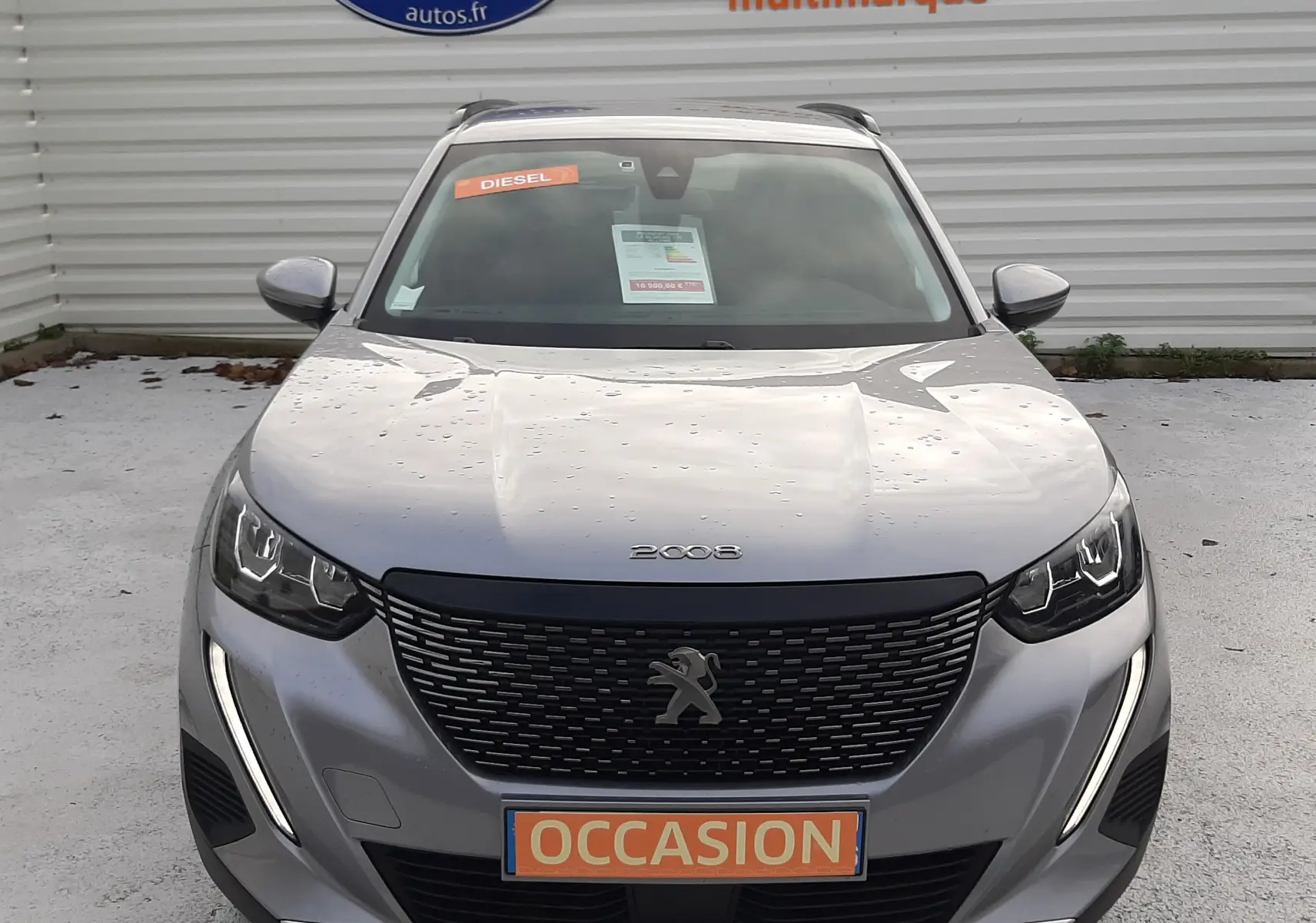 Vue frontale d'un Peugeot 2008 gris 2021 avec calandre noire et plaque "OCCASION" orange visible sur un parking.