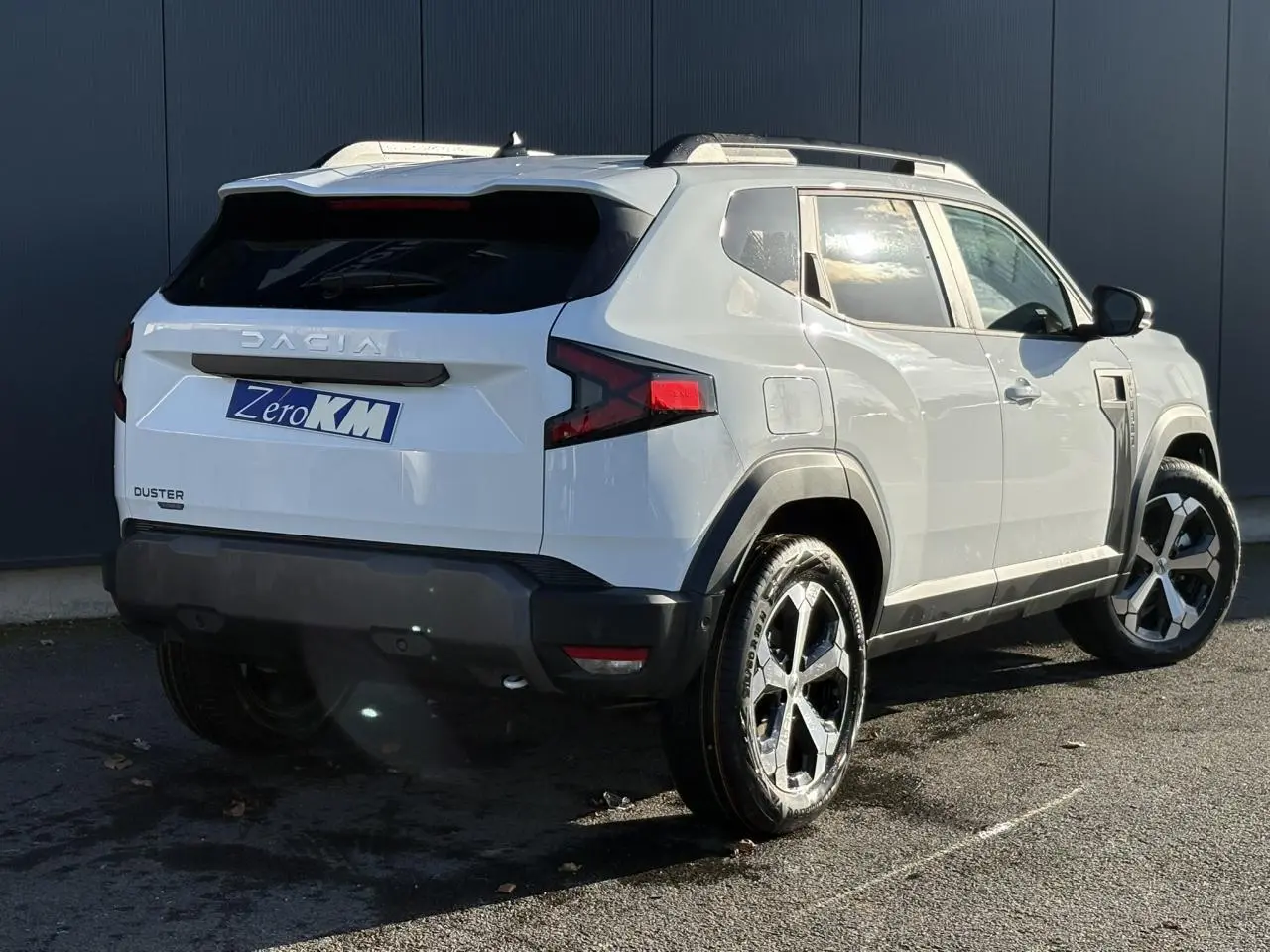 Vue 3/4 arrière droite du Dacia Duster blanc Glacier 2026 avec jantes alliage et barres de toit noires.