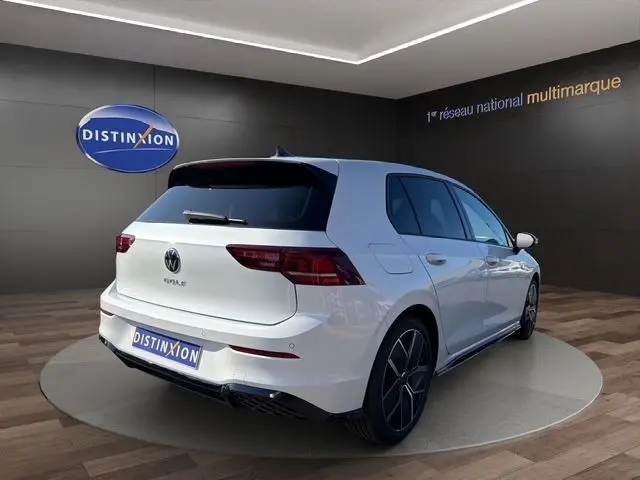 Vue 3/4 arrière droite d'une Volkswagen Golf blanche 2025 avec toit panoramique et jantes noires en showroom.