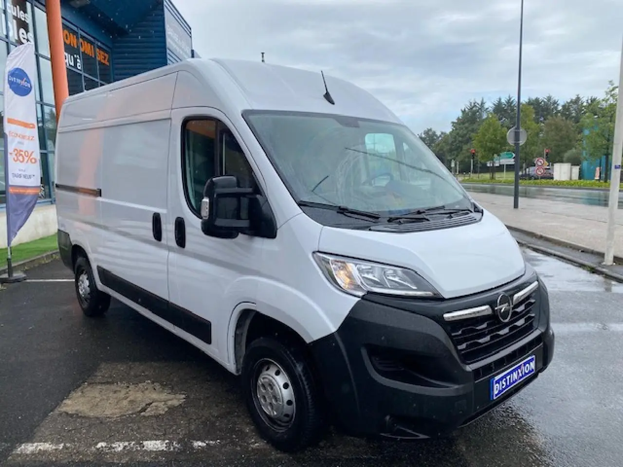Opel Movano Fourgon blanc vu en 3/4 avant droit sur parking humide devant un bâtiment commercial.