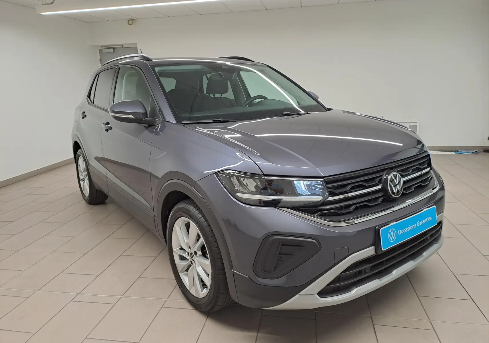 Volkswagen T-Cross gris cendré en 3/4 avant droit, avec calandre noire et jantes alliage visibles en intérieur showroom.
