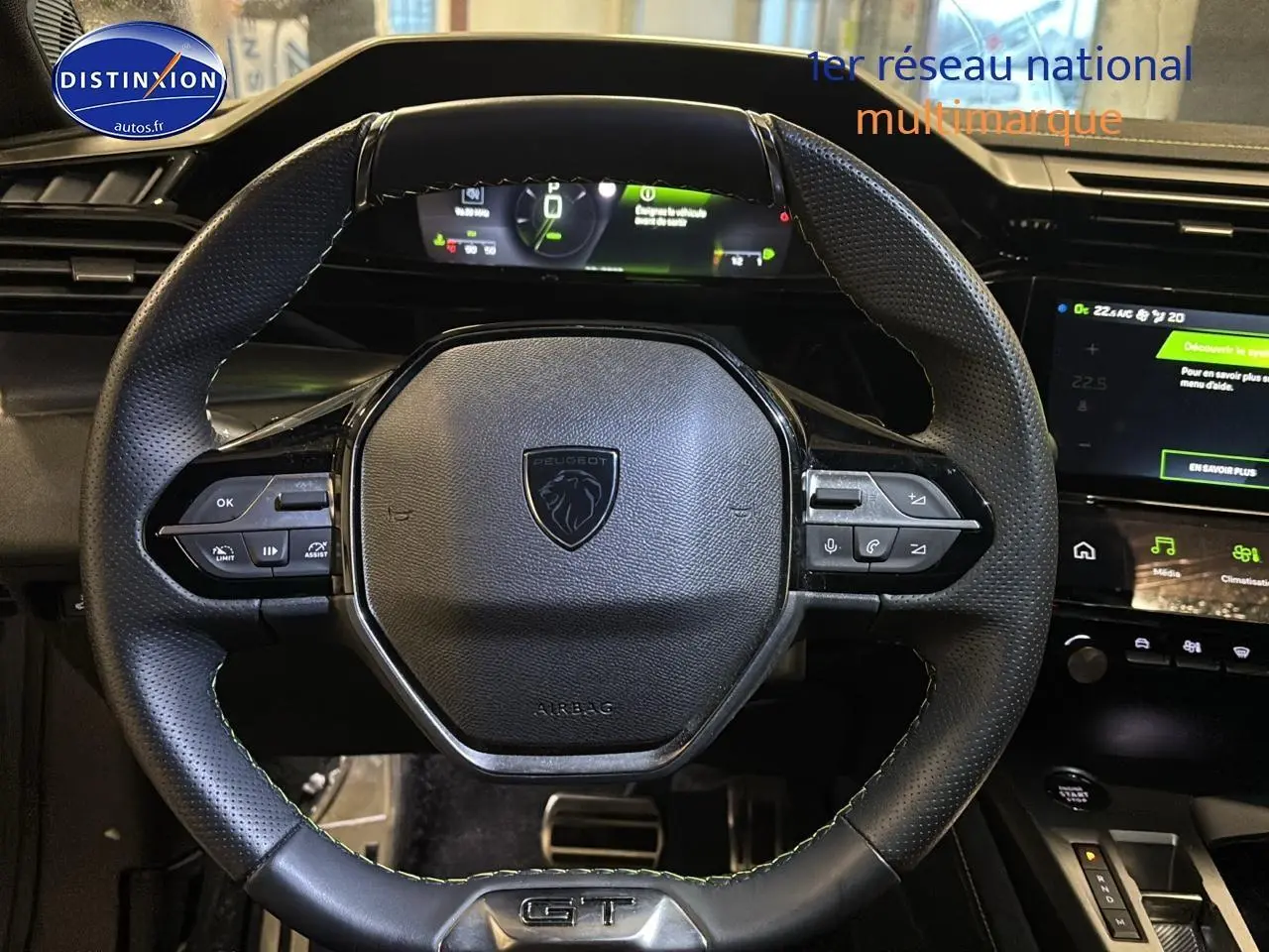 Vue rapprochée du volant cuir noir perforé avec logo Peugeot et badge GT, tableau de bord numérique en arrière-plan.