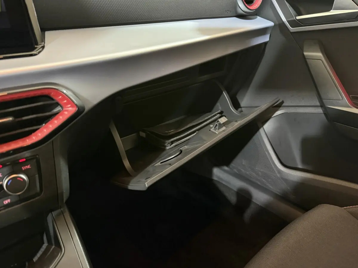 Intérieur de la SEAT Ibiza 2024 noir, vue sur la boîte à gants ouverte avec un livret à l’intérieur et détails rouges autour des aérateurs.
