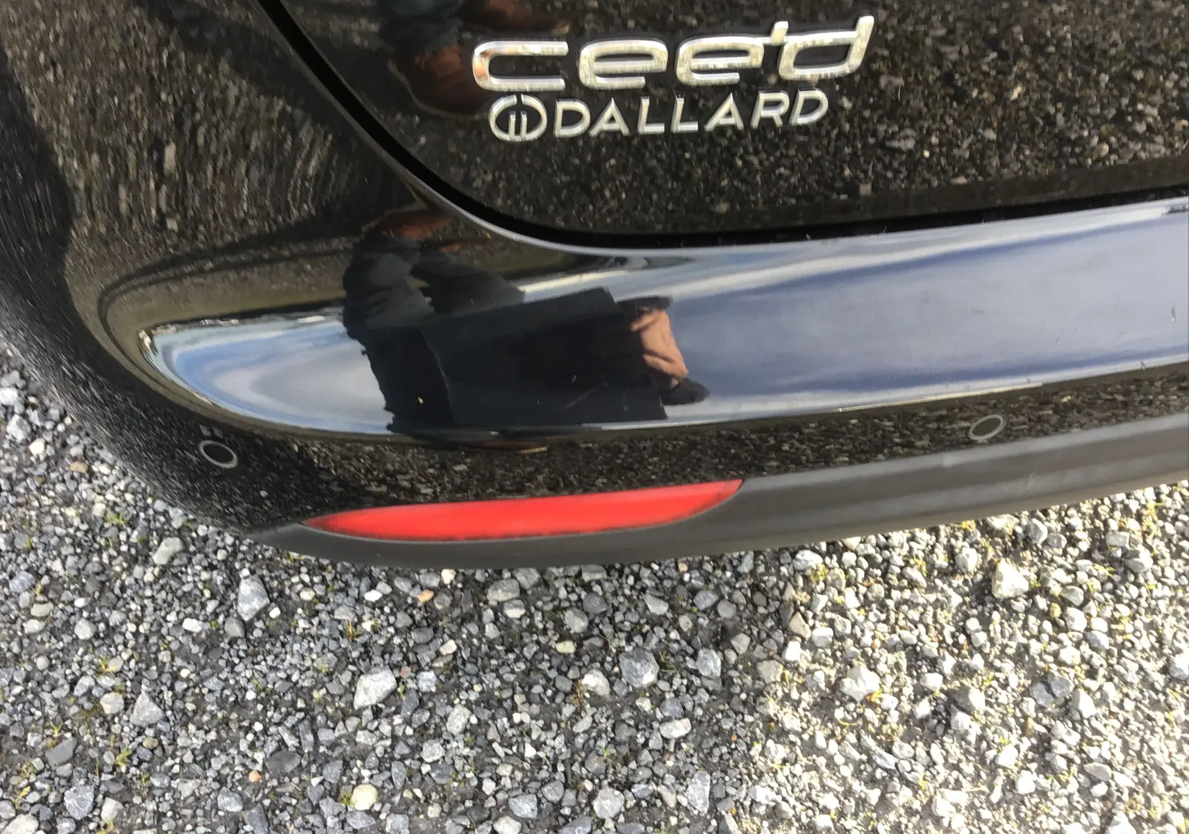 Gros plan sur le logo arrière et le pare-chocs noir brillant d'une KIA CEE'D SW 2017, avec reflet et sol gravillonné.