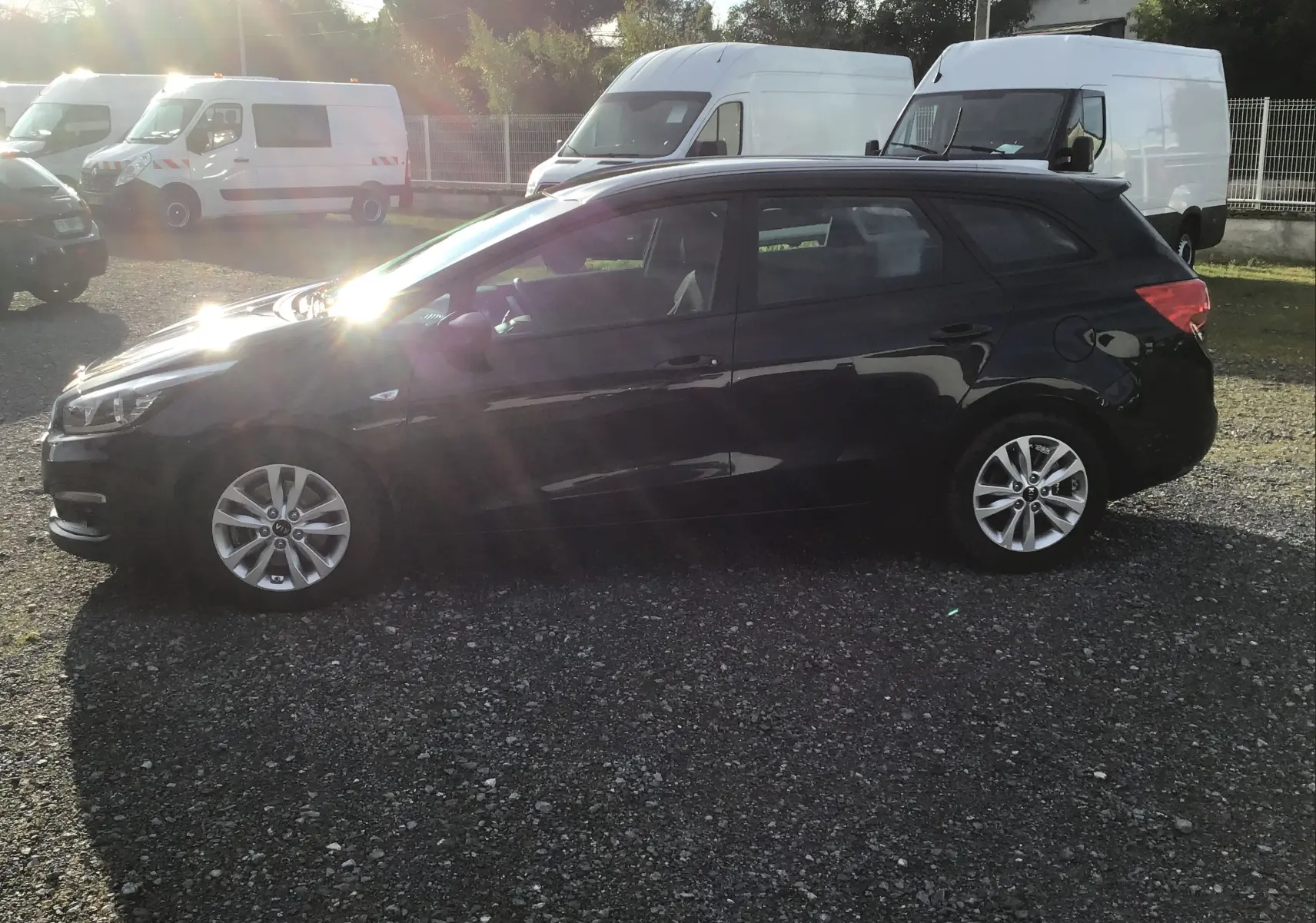Vue de profil côté gauche d'une Kia CEE'D SW noire 2017 garée sur un sol gravillonné avec des camionnettes blanches en arrière-plan.