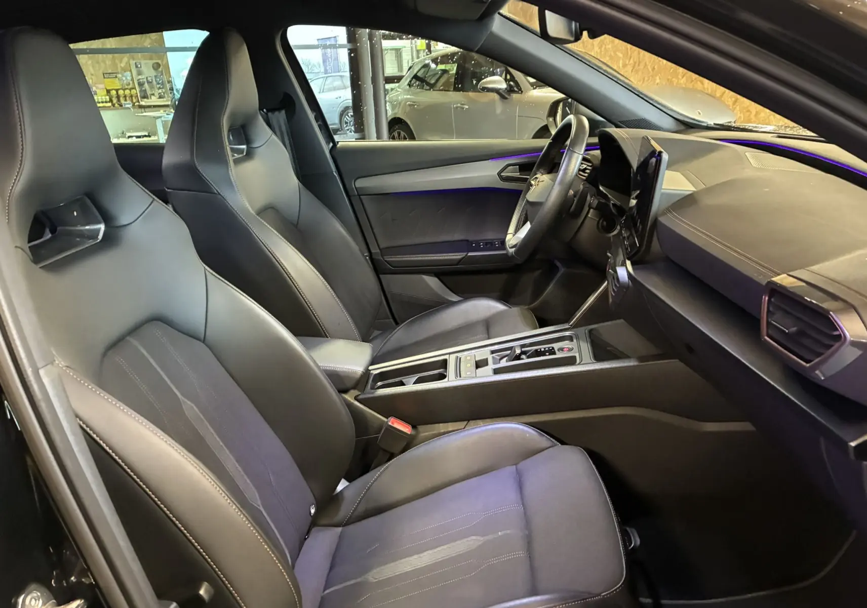 Vue intérieure côté conducteur du CUPRA Formentor noir 2023, sièges sport en tissu noir et tableau de bord moderne avec éclairage d’ambiance violet.