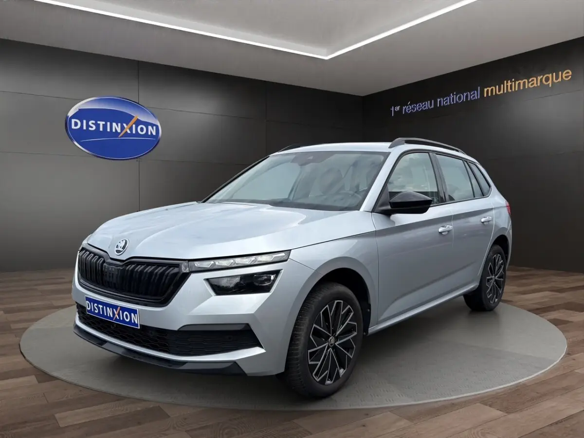 Vue 3/4 avant d'un Skoda Kamiq gris clair avec jantes bi-ton et rétroviseurs noirs dans un showroom.