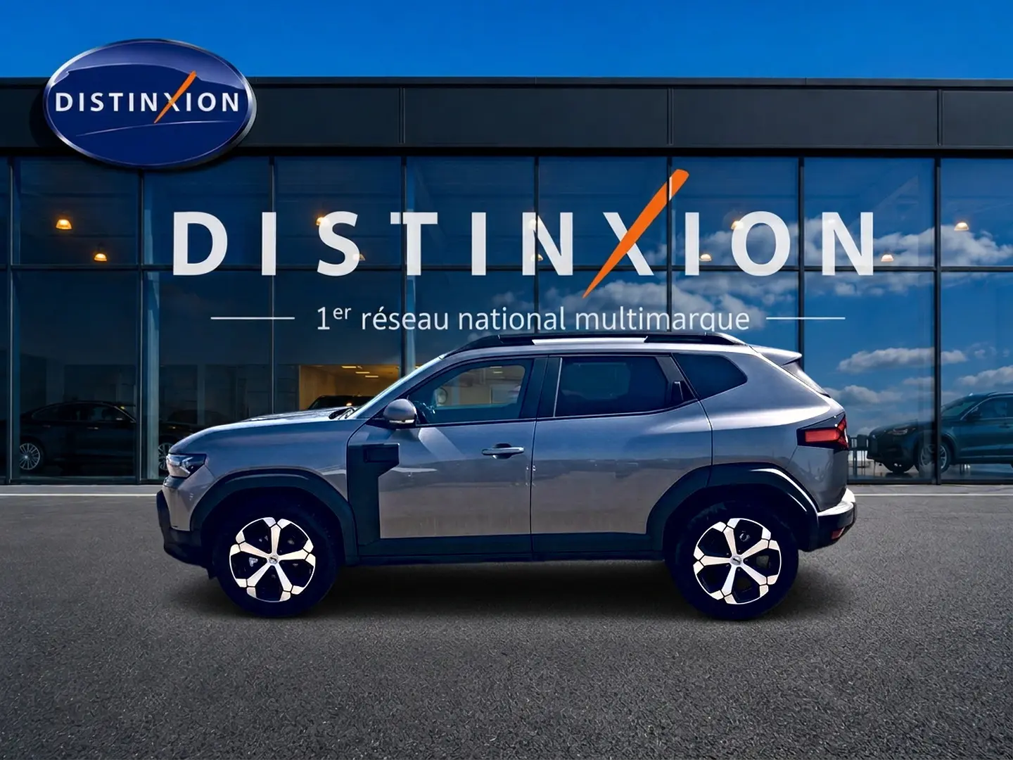 Profil droit du Dacia Duster gris schiste 2026 avec jantes alu et barres de toit visibles devant une vitrine Distinxion.