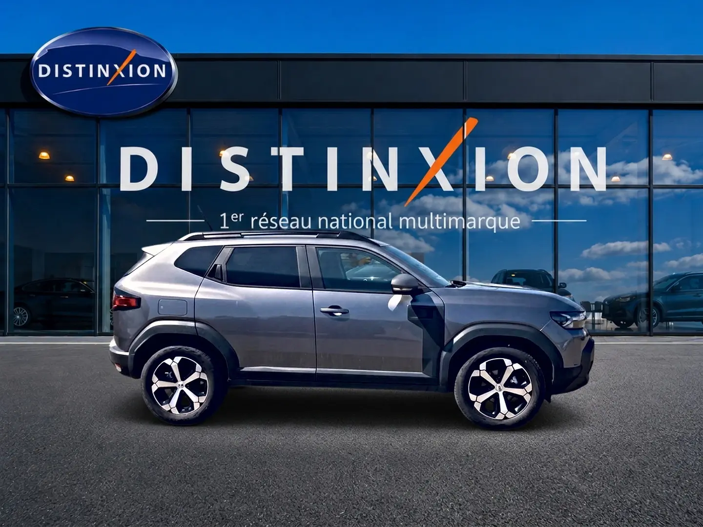 Vue de profil côté gauche d'un Dacia Duster gris schiste 2026 avec jantes alu et barres de toit, devant une vitrine Distinxion.