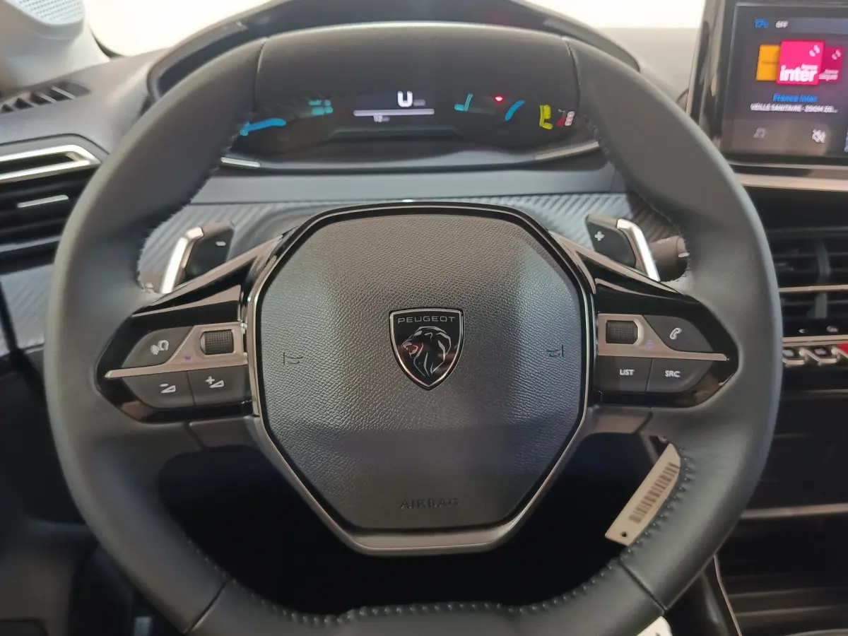 Vue rapprochée du volant cuir multifonctions de la Peugeot 208 Hybrid gris clair avec tableau de bord numérique en arrière-plan.