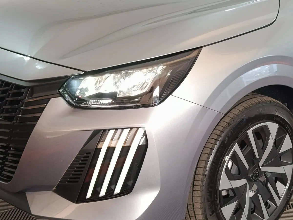 Gros plan sur l'avant droit gris clair d'une Peugeot 208 Hybrid 100 e-DCS6 avec phares LED et jante stylisée.