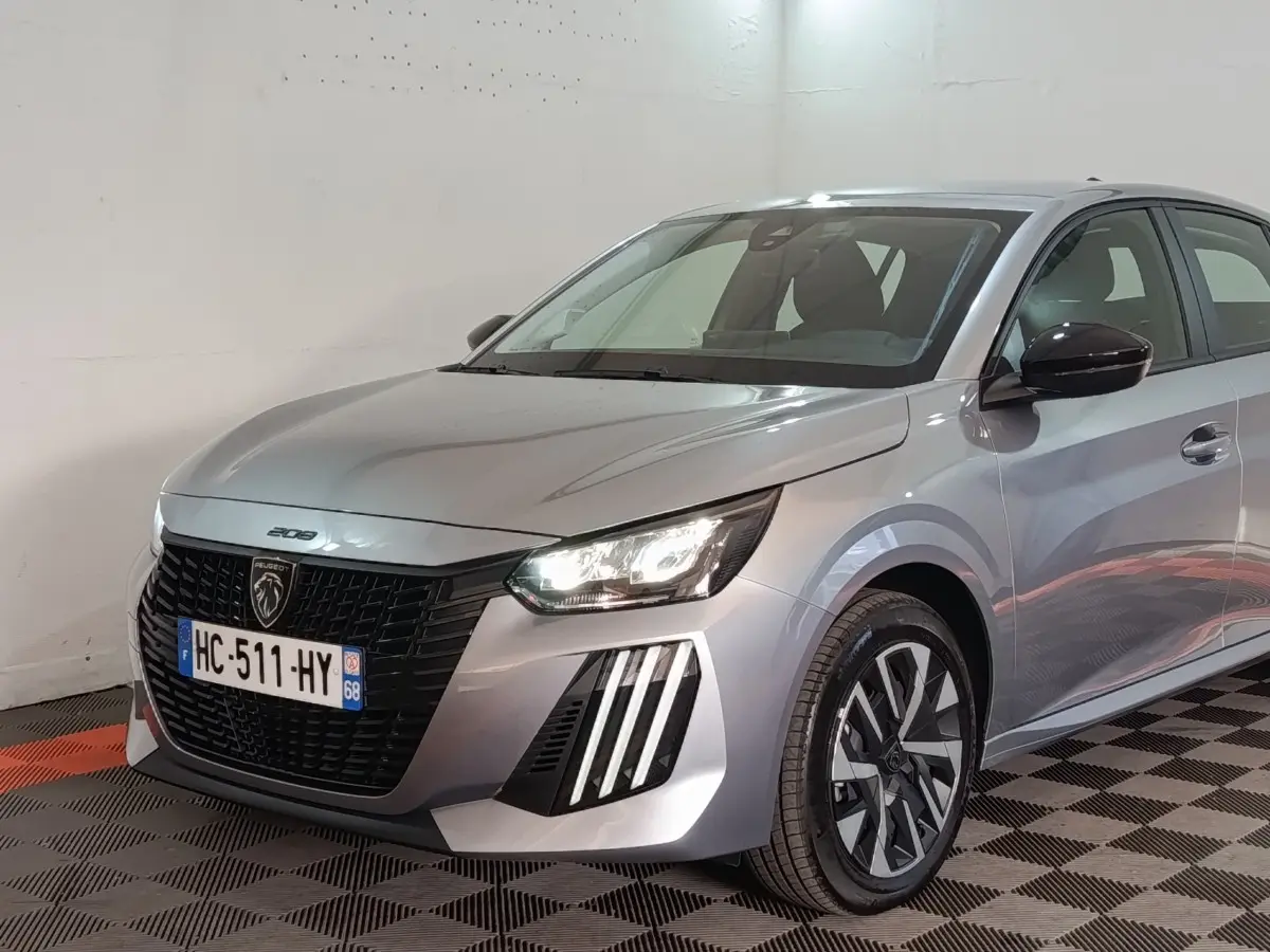 Peugeot 208 Hybrid gris clair vue 3/4 avant droit, phares LED allumés et jantes distinctives bicolores.