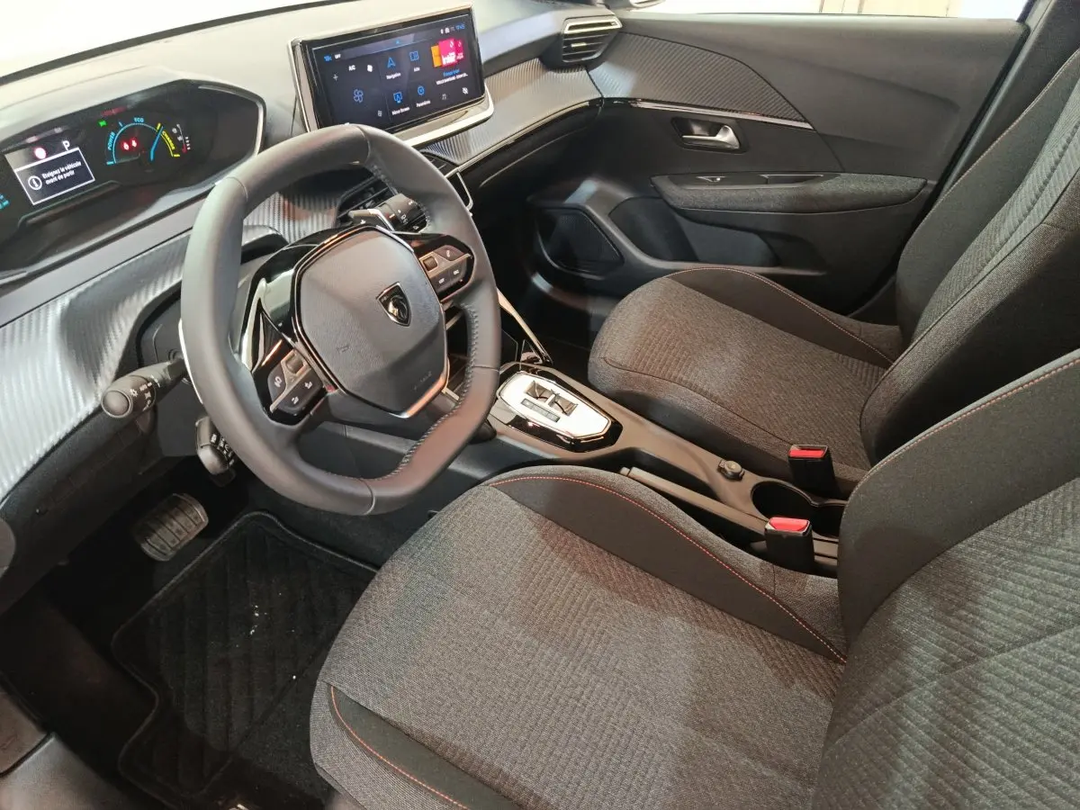 Vue intérieure côté conducteur de la Peugeot 208 Hybrid 2025, volant cuir multifonctions et sièges tissu gris avec surpiqûres rouges.