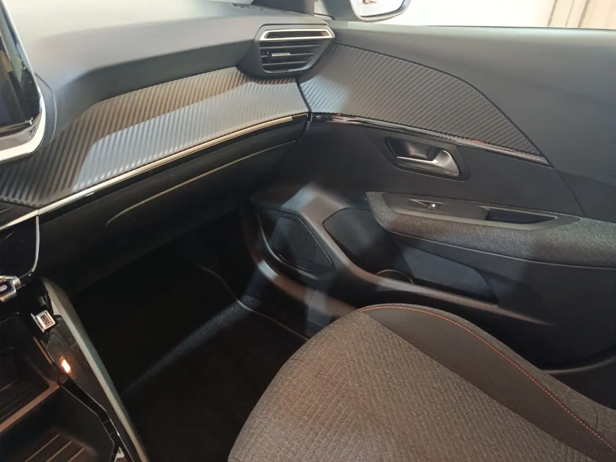 Vue intérieure côté passager de la Peugeot 208 Hybrid 2025, tableau de bord avec finition carbone et siège tissu gris.