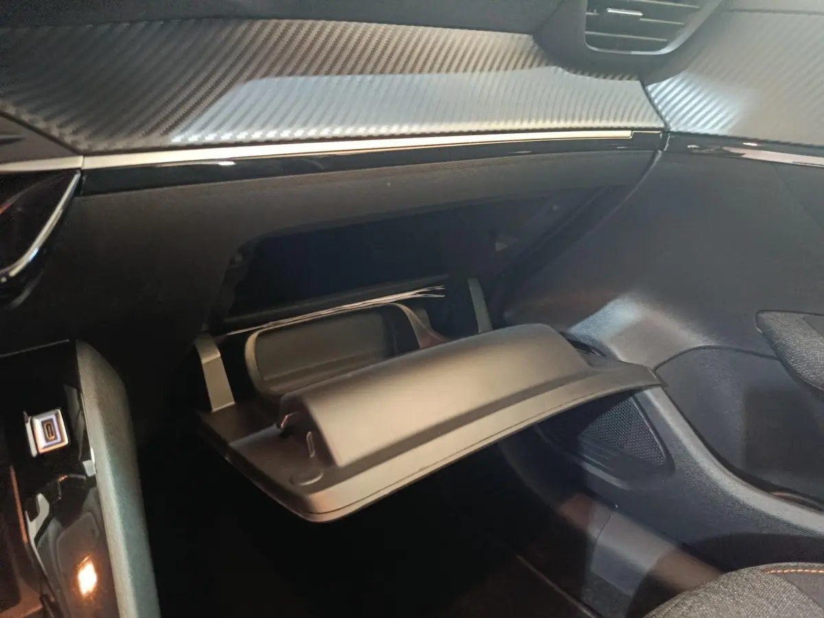 Vue intérieure du coffre à gants ouvert sur le côté passager d’une Peugeot 208 Hybrid gris clair, tableau de bord avec finition carbone.