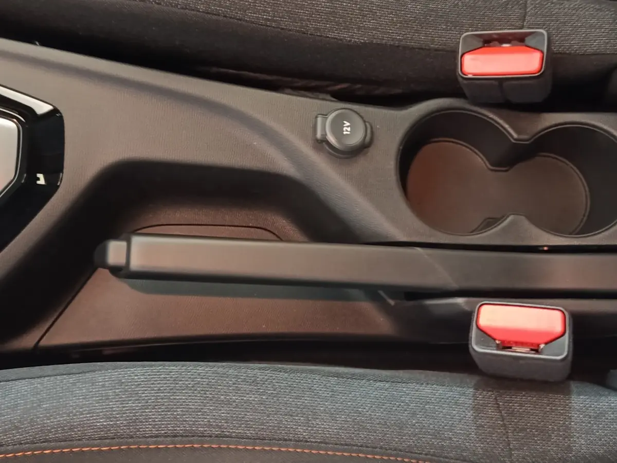 Vue rapprochée de la console centrale de la Peugeot 208 Hybrid 2025, avec frein à main, prise 12V et porte-gobelets noirs.