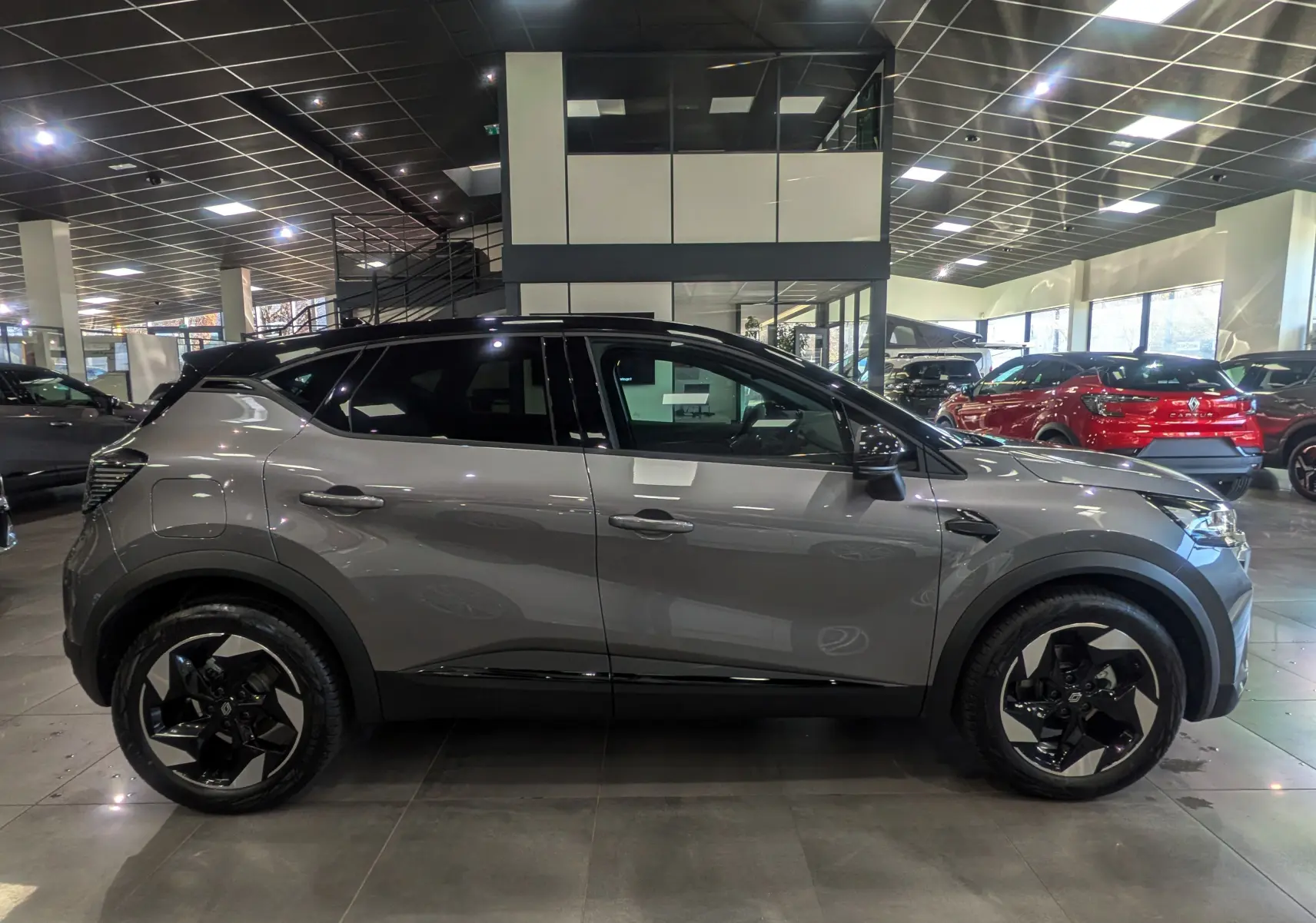 Profil côté gauche du Renault Captur 2025 gris Cassiopé avec toit noir et jantes biton dans un showroom.