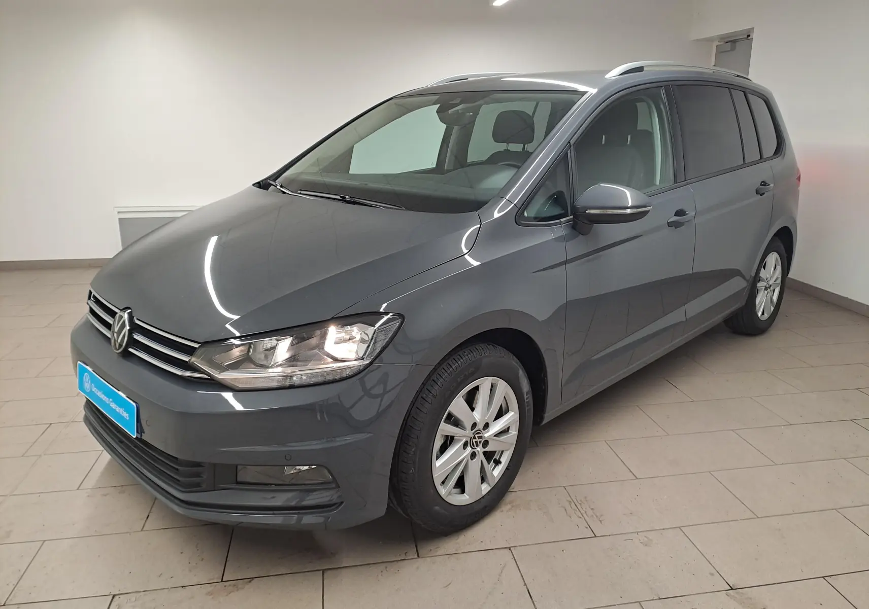 Volkswagen Touran gris dauphin en 3/4 avant droit, avec jantes alliage et phares allumés, intérieur visible.