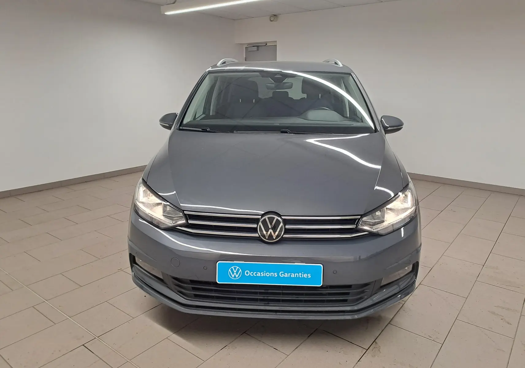 Vue avant d'un Volkswagen Touran gris dauphin 2024 avec phares allumés dans un showroom lumineux.