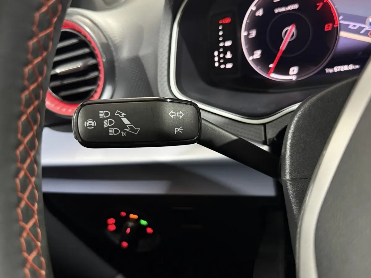 Gros plan sur la manette de clignotants noire avec icônes blanches, volant cuir gris foncé avec surpiqûres rouges, tableau de bord digital.