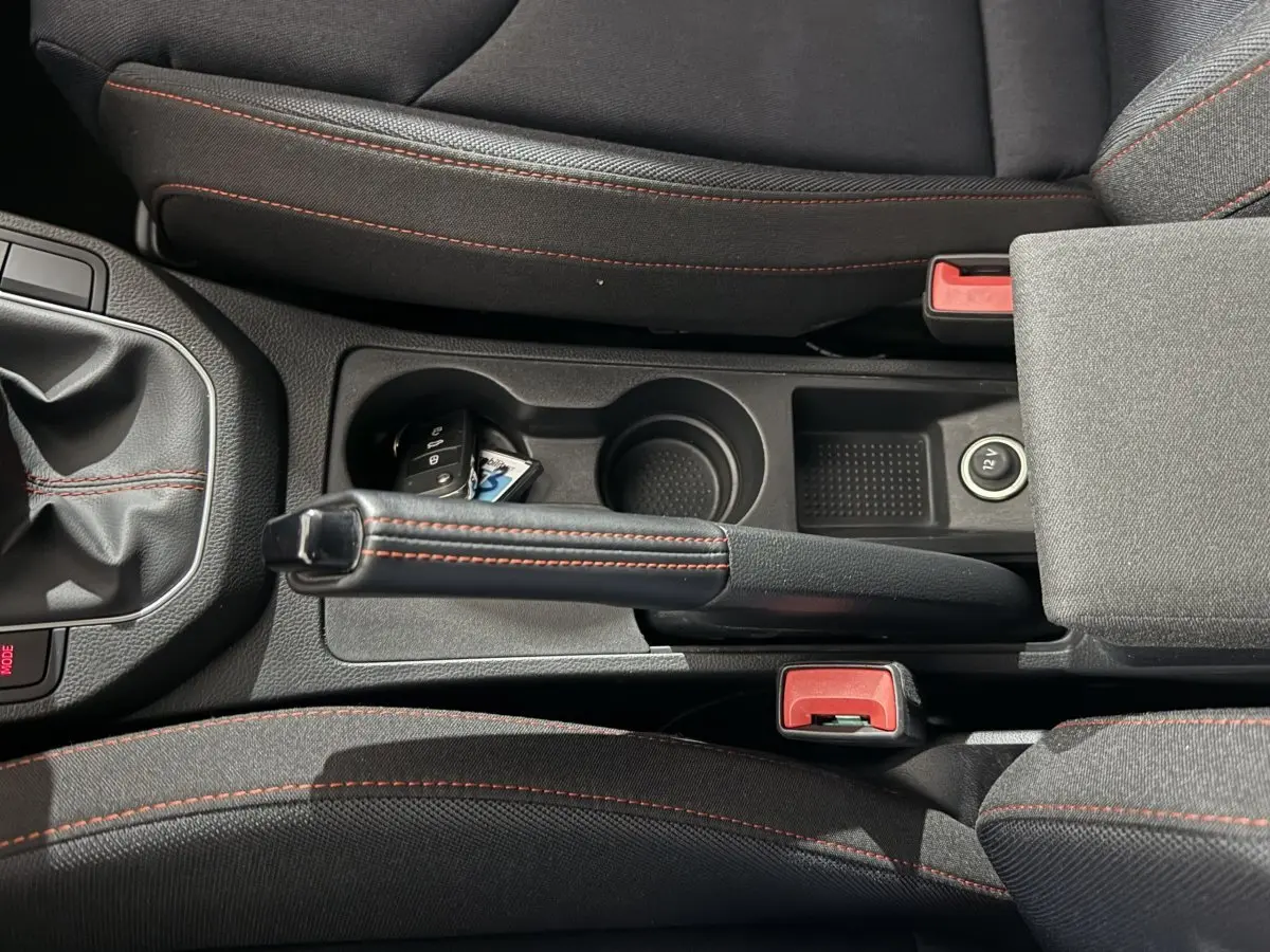 Vue plongeante sur la console centrale noire avec frein à main cuir et coutures rouges, clé et prise 12V visibles.