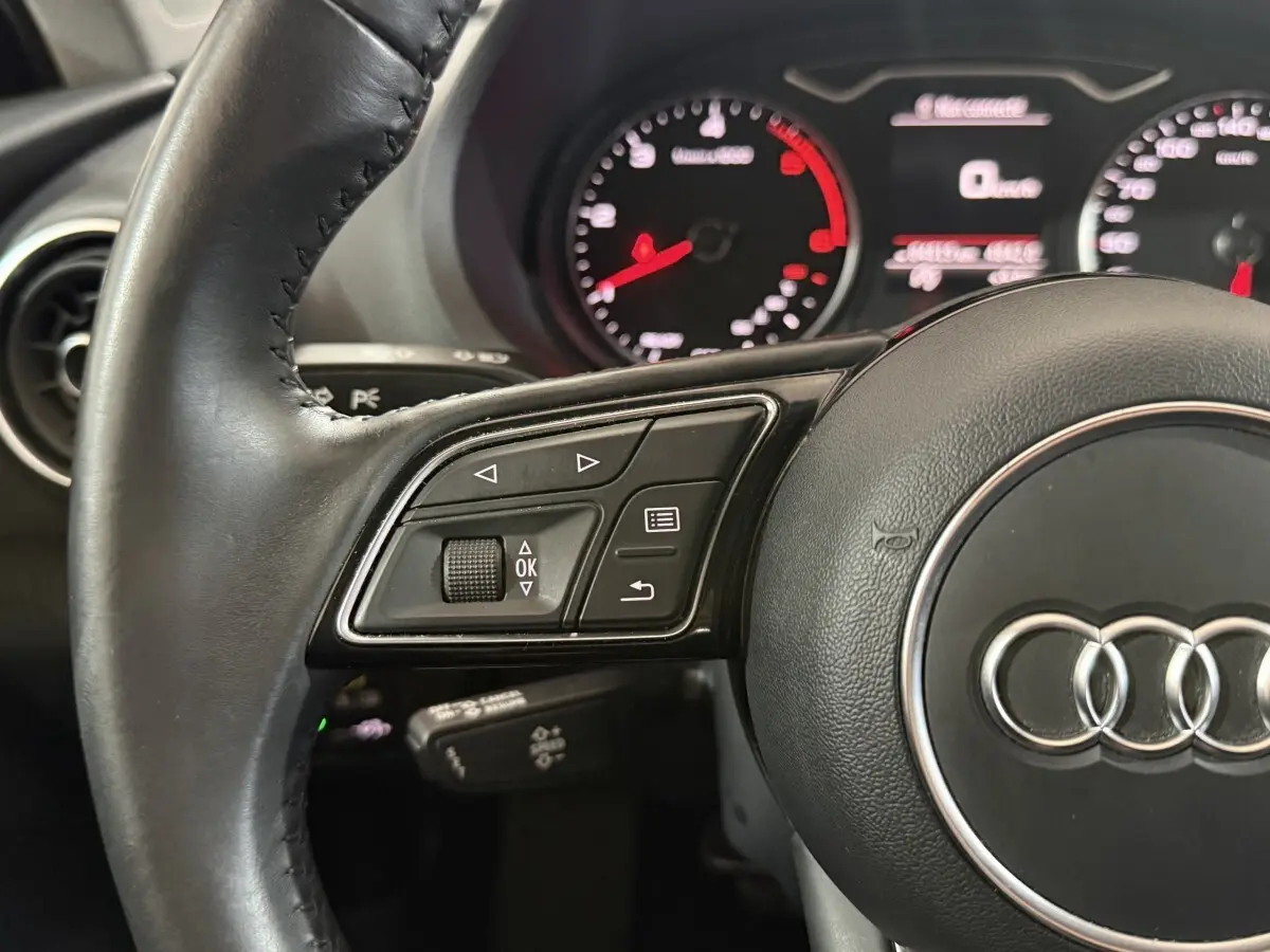 Vue rapprochée du volant cuir noir de l'Audi A3 2018 avec commandes multifonctions et tableau de bord éclairé.