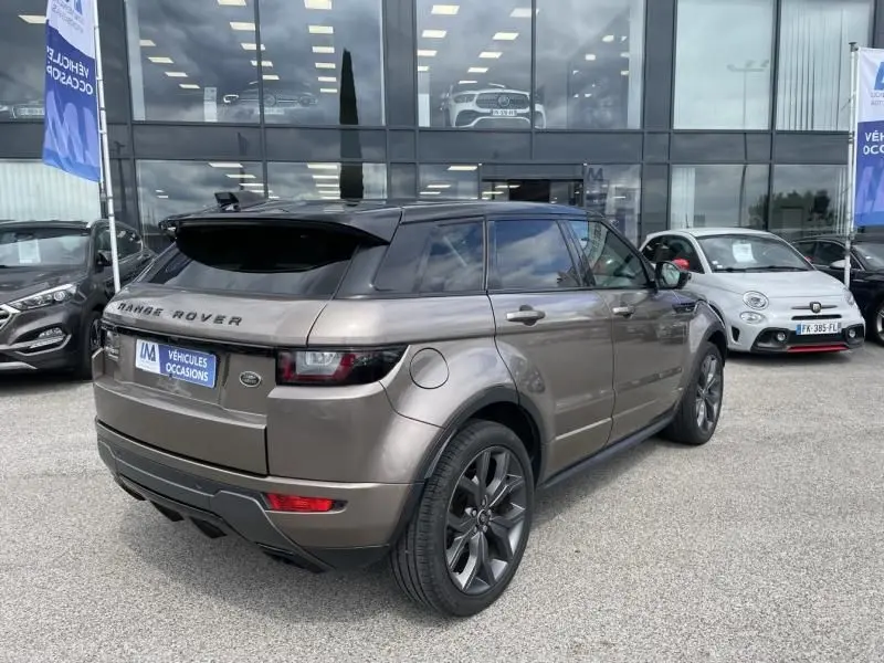 Vue 3/4 arrière droite d'un Range Rover Evoque 2017 marron métal avec jantes foncées, stationné devant un showroom.