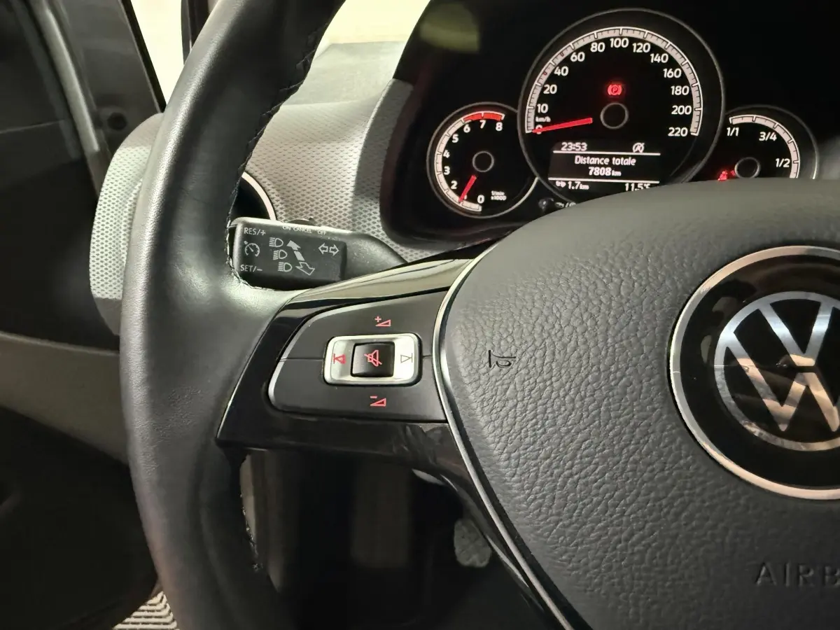 Gros plan sur le volant cuir noir de la Volkswagen Up 2022, avec commandes audio et tableau de bord éclairé en arrière-plan.
