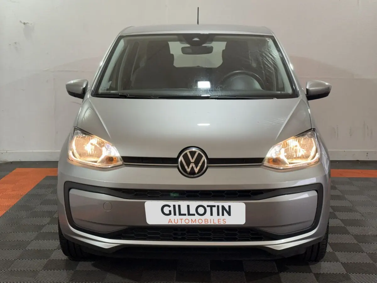 Vue avant d'une Volkswagen Up gris foncé avec phares allumés sur sol intérieur à damier noir et orange.