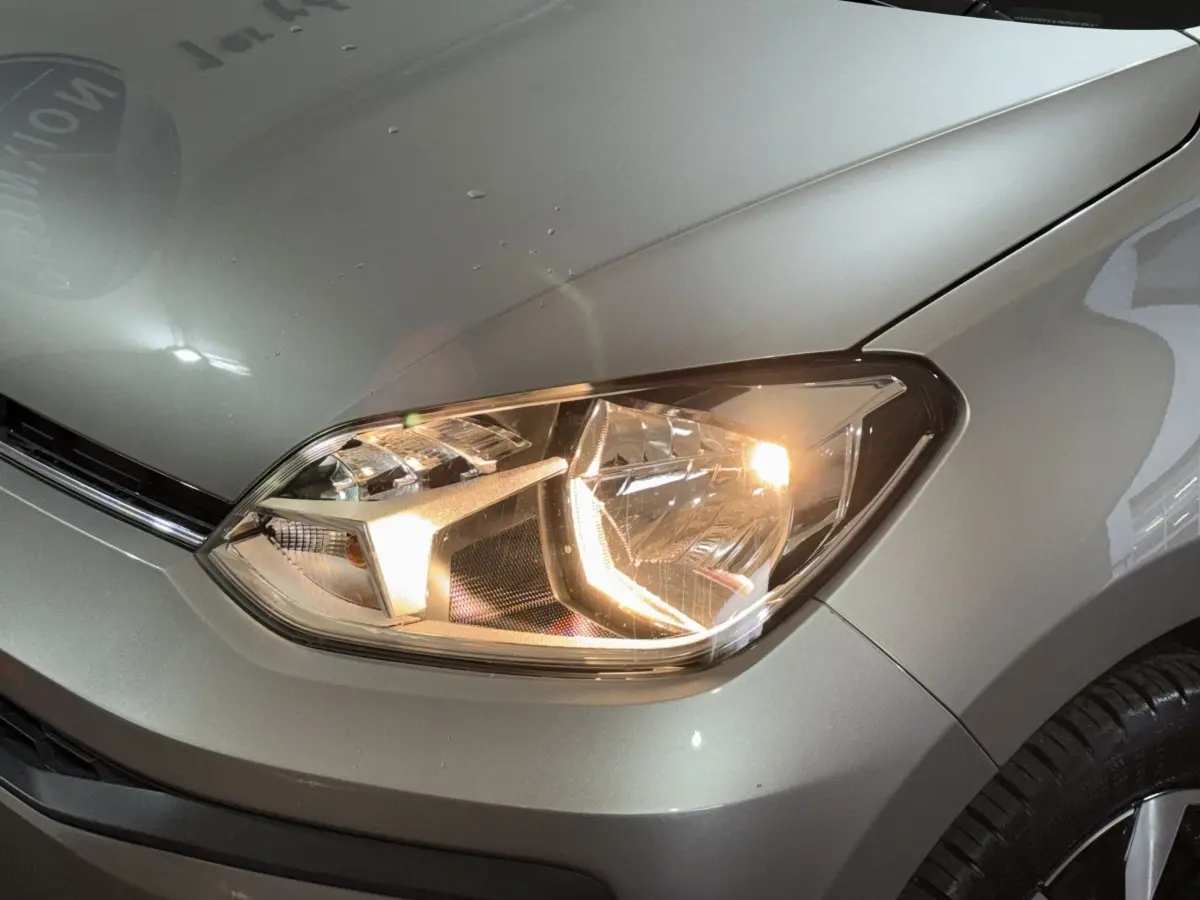 Gros plan sur le phare avant droit allumé d'une Volkswagen Up gris foncé, avec partie du capot visible.