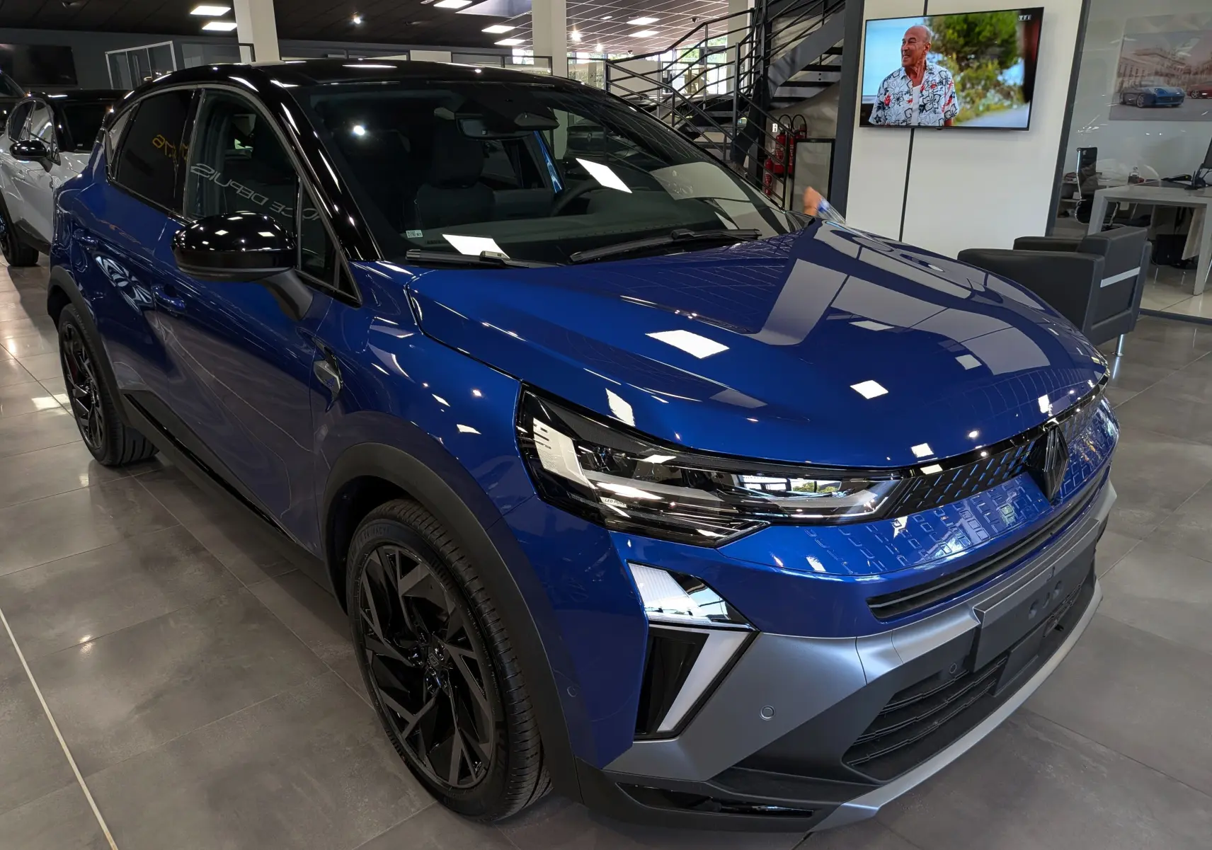 Renault Captur E-Tech hybride 2026 bleu Iron avec toit noir, vue 3/4 avant droit en intérieur showroom.
